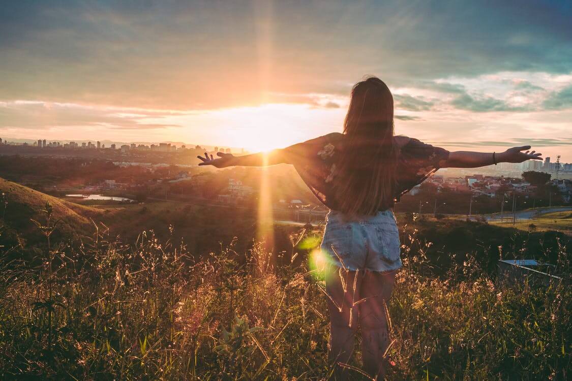 Woman with arms outstretched looking out at a city from atop a grassy hill at sunset