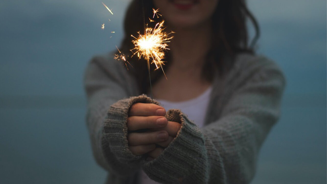 Woman wearing a long sleeved sweater celebrating with a sparkler glowing in her hands