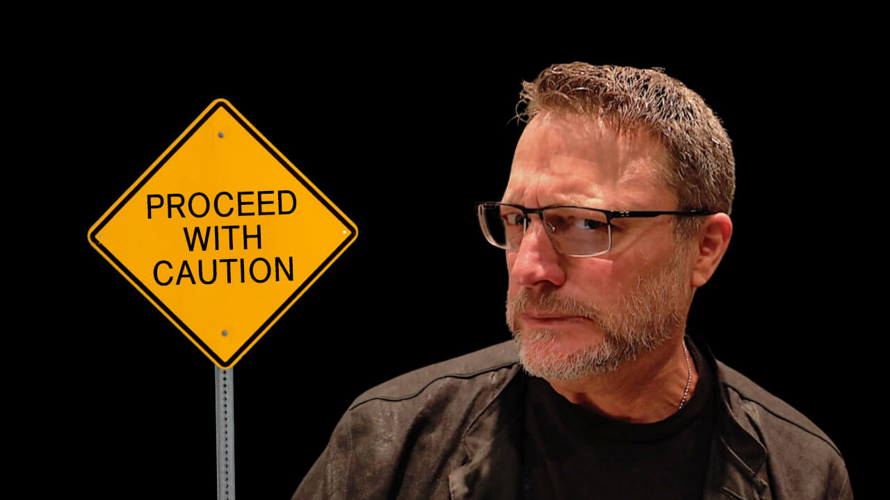 Steve Blum making a silly face near a yellow Proceed With Caution sign in front of a black background